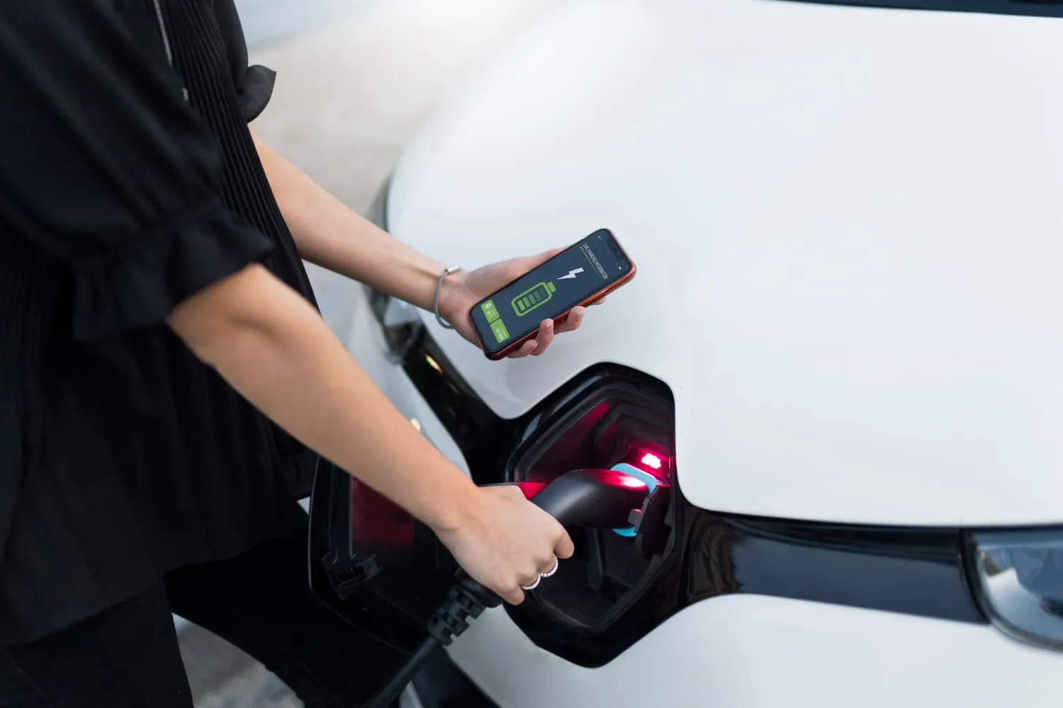 一個人正將電動車接上充電插頭，並用手機查看充電狀態，螢幕上顯示電池圖示與充電進度。